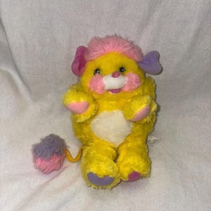 May include: A plush toy featuring a bright yellow body with a white belly patch, pink cheek fur, and a pink tuft of hair on its head. It has one pink ear, one purple ear, and a long tail ending in a fluffy purple and pink pom-pom ball.