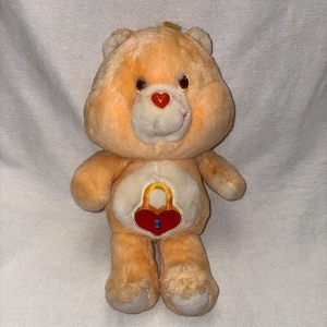May include: A plush, peach-colored Care Bear with a heart-shaped nose and a red heart-shaped lock on its belly. The bear has a white muzzle and brown eyes. The lock has a gold-colored lock and a blue keyhole.