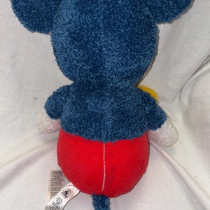May include: A plush Mickey Mouse toy, viewed from the back. The toy features large, dark blue ears and a matching body. It has red corduroy shorts, white gloves, and a blue tail. A yellow accent is visible.