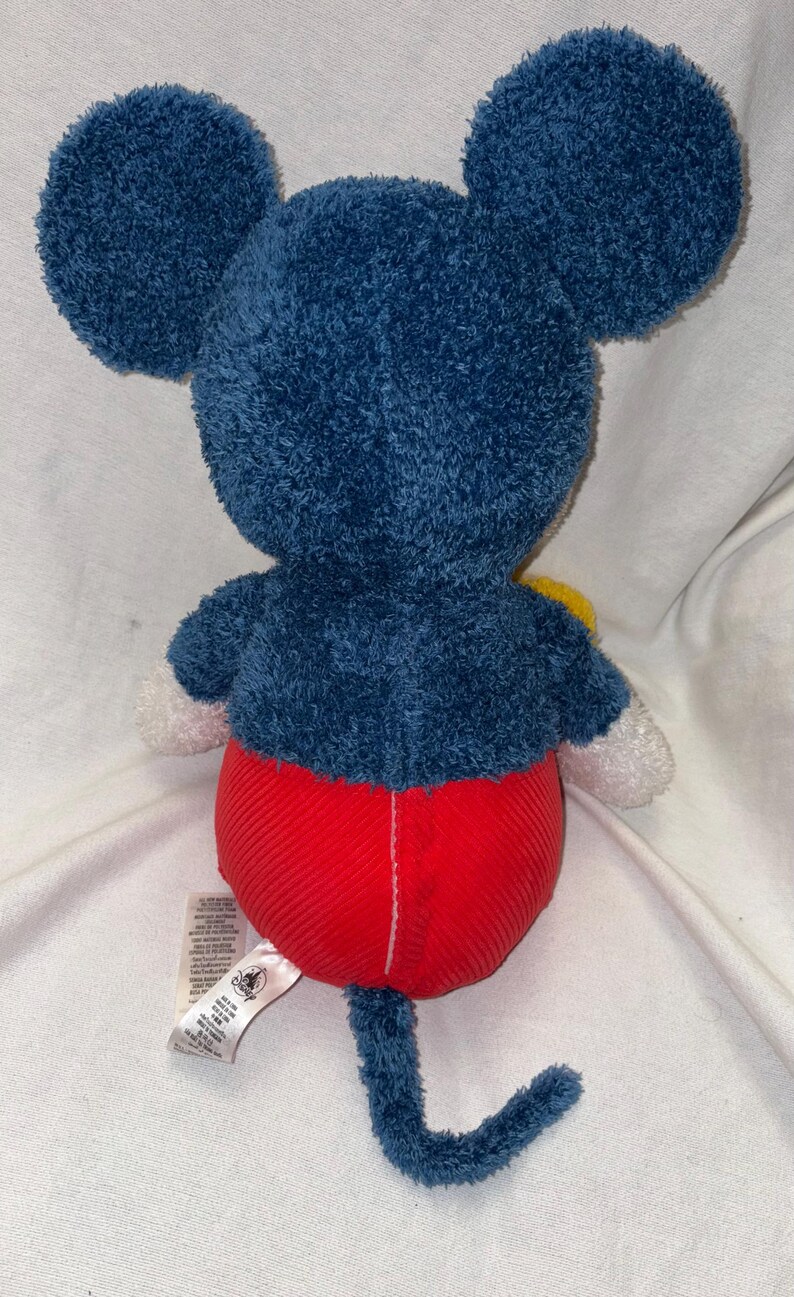 May include: A plush Mickey Mouse toy, viewed from the back. The toy features large, dark blue ears and a matching body. It has red corduroy shorts, white gloves, and a blue tail. A yellow accent is visible.