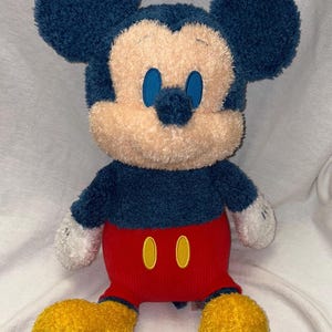 May include: A plush Mickey Mouse toy with a navy blue head and ears, a tan face, and blue eyes. It wears a red pair of shorts with two yellow buttons and yellow feet. The toy has white gloves and is sitting upright.