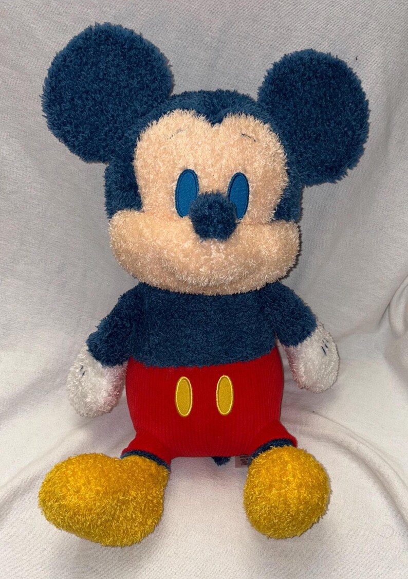 May include: A plush Mickey Mouse toy with a navy blue head and ears, a tan face, and blue eyes. It wears a red pair of shorts with two yellow buttons and yellow feet. The toy has white gloves and is sitting upright.