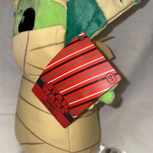 Puede incluir: Un peluche de Grogu de Star Wars, con cabeza y orejas verdes, y un atuendo de envoltura beige. El peluche tiene una etiqueta roja con rayas negras y blancas y el logo de Star Wars.