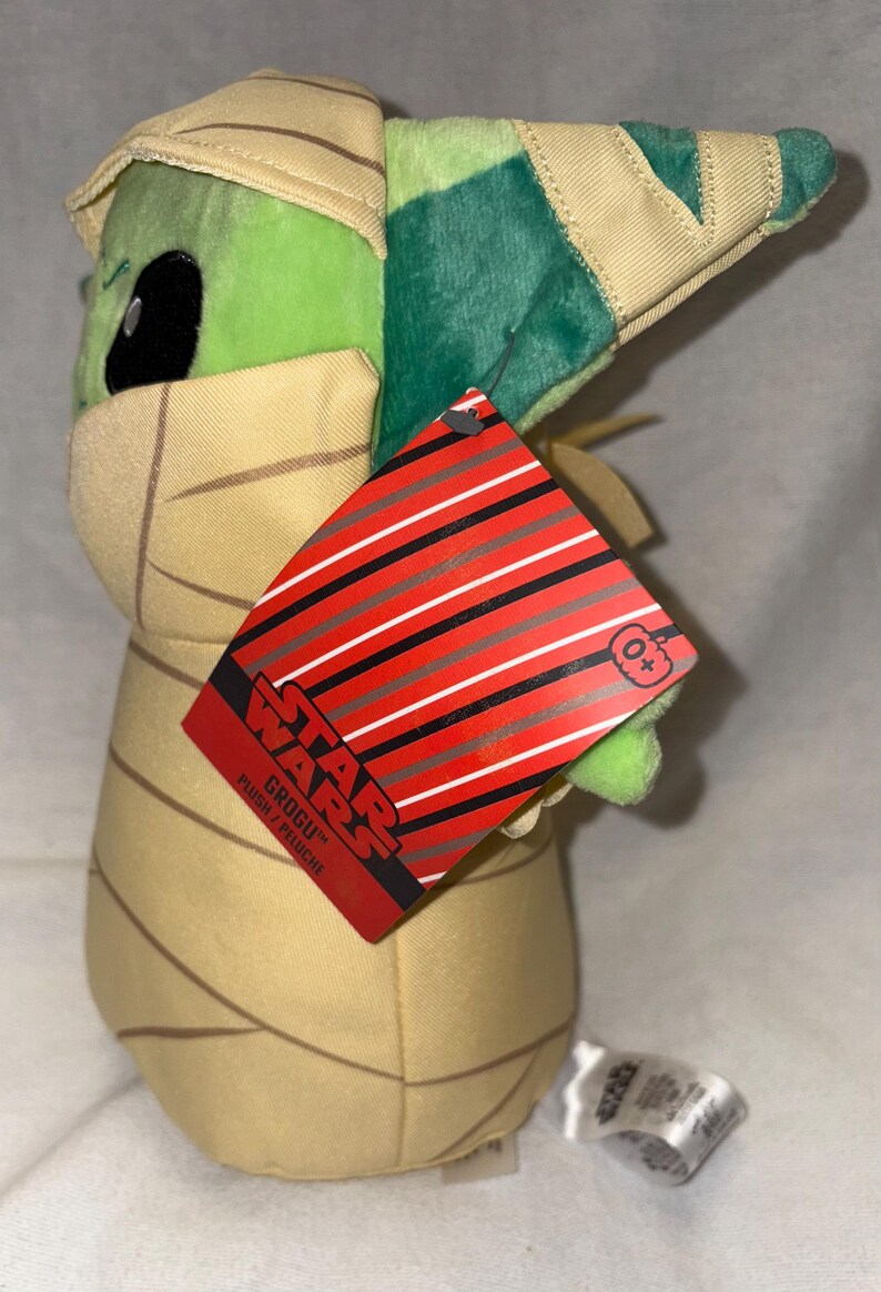 Puede incluir: Un peluche de Grogu de Star Wars, con cabeza y orejas verdes, y un atuendo de envoltura beige. El peluche tiene una etiqueta roja con rayas negras y blancas y el logo de Star Wars.