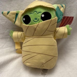 Puede incluir: Un peluche de un personaje alien&iacute;gena verde envuelto en vendas color beige, que se asemeja a una momia. El juguete tiene grandes ojos negros, una peque&ntilde;a sonrisa y orejas grandes. El juguete sostiene un peque&ntilde;o objeto rojo y marr&oacute;n.