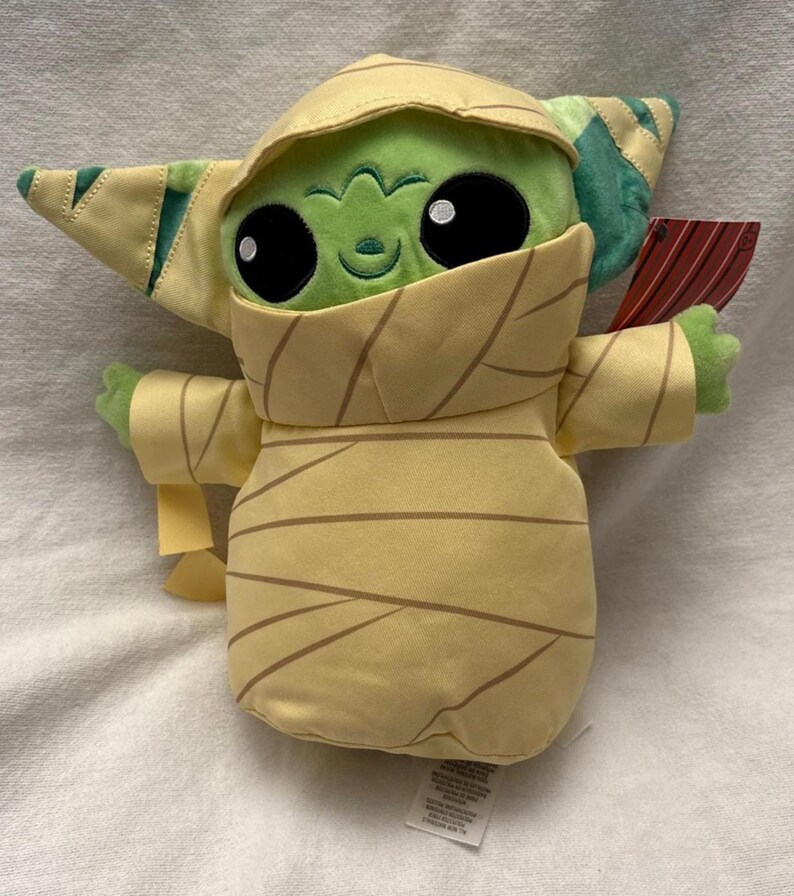 Puede incluir: Un peluche de un personaje alien&iacute;gena verde envuelto en vendas color beige, que se asemeja a una momia. El juguete tiene grandes ojos negros, una peque&ntilde;a sonrisa y orejas grandes. El juguete sostiene un peque&ntilde;o objeto rojo y marr&oacute;n.