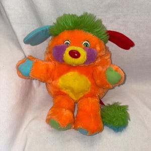 May include: A plush toy featuring a bright orange body with a yellow heart-shaped patch on the chest. It has green hair, one blue ear, one red ear, purple cheek patches, a red nose, and green and blue accents on the paws and tail.