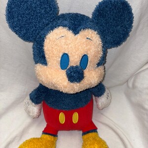 May include: A plush Mickey Mouse toy with a blue head and ears, a tan face, and blue eyes. It has a red body with two yellow buttons and yellow feet. The toy is soft and cuddly.