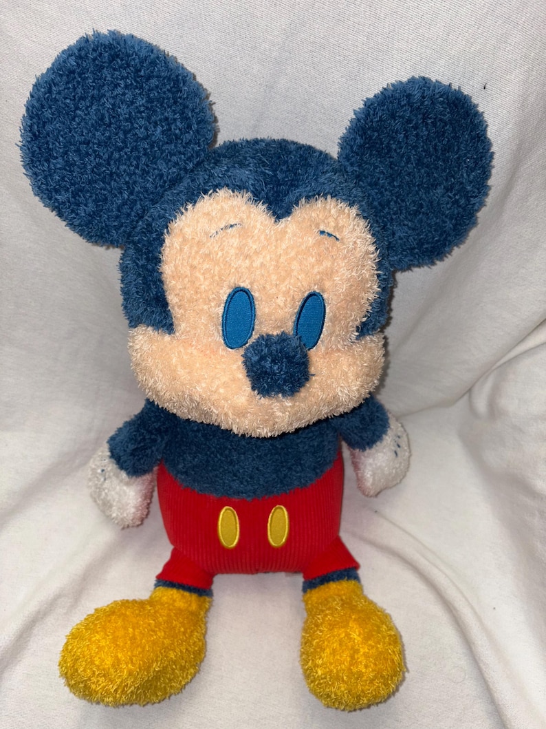 May include: A plush Mickey Mouse toy with a blue head and ears, a tan face, and blue eyes. It has a red body with two yellow buttons and yellow feet. The toy is soft and cuddly.