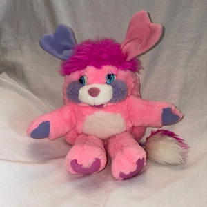 May include: A plush toy creature with bright pink fur, a tuft of magenta hair, and large blue eyes. It features one purple ear and one pink ear, purple cheek patches, a white muzzle with a small nose, and purple paw pads. It has a fluffy white tail.