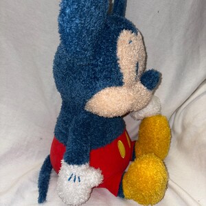 May include: A plush Mickey Mouse toy in a seated position. The toy features a blue head and ears, a tan face, red shorts, yellow feet, and white gloves with embroidered details. A classic Disney character.