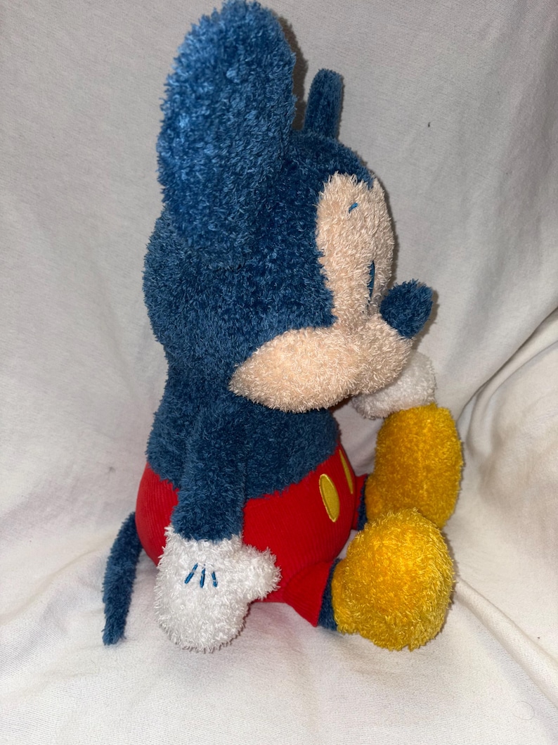 May include: A plush Mickey Mouse toy in a seated position. The toy features a blue head and ears, a tan face, red shorts, yellow feet, and white gloves with embroidered details. A classic Disney character.