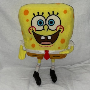 May include: Yellow plush toy of SpongeBob SquarePants with a red tie and black shoes. He is smiling with his teeth showing.