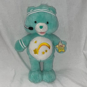 May include: A teal blue Care Bear plush toy wearing a white headband and a headset. The bear has a rainbow and stars on its chest and a tag that reads "At Home. My music moves me and helps me work out in 3 fun ways."