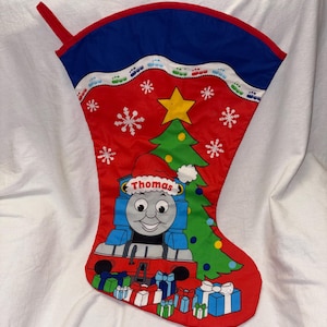 May include: A red Christmas stocking featuring a graphic of Thomas the Tank Engine wearing a Santa hat. The design includes a green Christmas tree with a yellow star, white snowflakes, and colorful gift boxes at the base. A blue cuff tops the stocking.