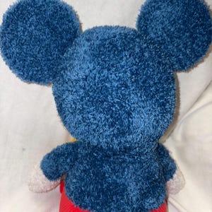 May include: A plush toy of a famous cartoon character, viewed from the back. The toy has large, round, blue ears and a matching blue body. The arms have white gloves, and the pants are red. The toy is made of a soft, textured material.