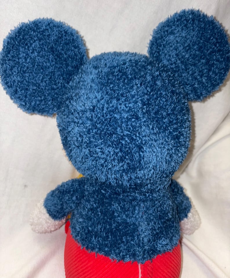 May include: A plush toy of a famous cartoon character, viewed from the back. The toy has large, round, blue ears and a matching blue body. The arms have white gloves, and the pants are red. The toy is made of a soft, textured material.
