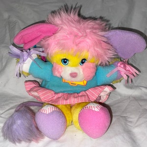 May include: A pink and yellow plush toy with a blue shirt and pink skirt. The toy has a long tail with purple and pink fur. The toy has a pink nose and blue eyes.