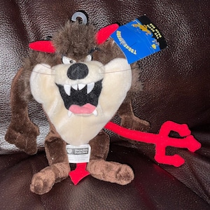May include: Brown plush Tazmanian Devil toy with red horns and a red tail with a trident. The toy is sitting on a brown surface. The tag reads "Toons Mania! Warner Bros. Studio Store"