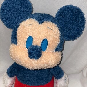 May include: A plush Mickey Mouse toy with a blue head and ears, a tan face, and blue eyes. The toy has a blue body, red corduroy pants with yellow buttons, and white gloves. The toy is soft and cuddly.