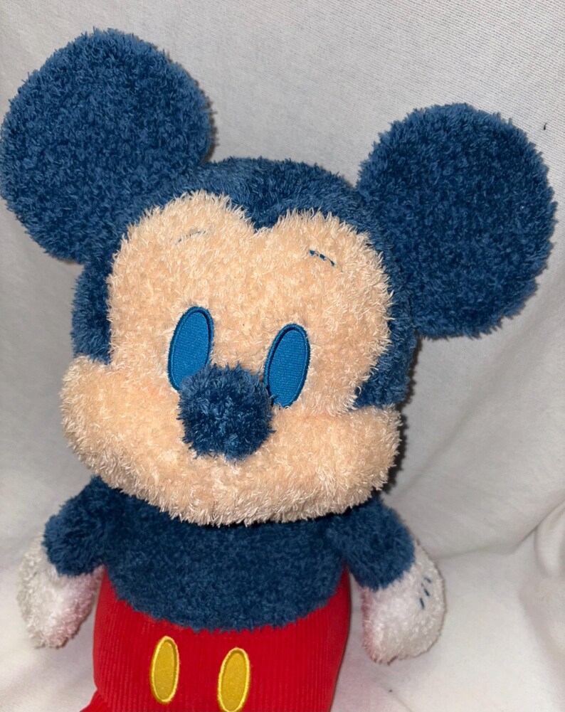 May include: A plush Mickey Mouse toy with a blue head and ears, a tan face, and blue eyes. The toy has a blue body, red corduroy pants with yellow buttons, and white gloves. The toy is soft and cuddly.