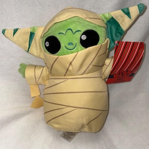 Puede incluir: Un peluche de un personaje de piel verde envuelto en vendas color beige, que se asemeja a una momia. El personaje tiene grandes ojos negros, una peque&ntilde;a sonrisa y orejas puntiagudas con detalles verdes y beige. Una etiqueta roja con rayas blancas y negras est&aacute; adjunta.