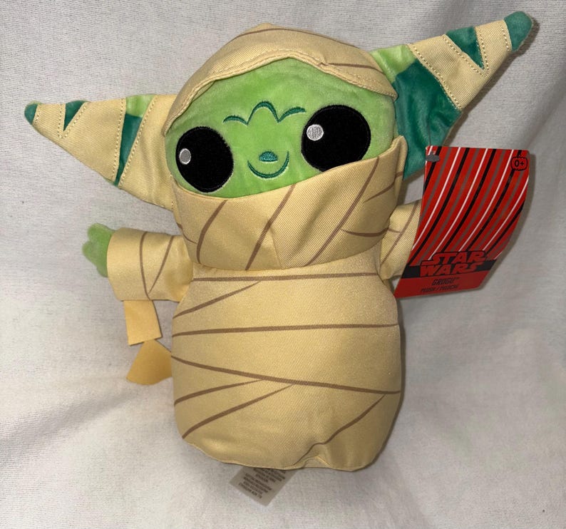 Puede incluir: Un peluche de un personaje de piel verde envuelto en vendas color beige, que se asemeja a una momia. El personaje tiene grandes ojos negros, una peque&ntilde;a sonrisa y orejas puntiagudas con detalles verdes y beige. Una etiqueta roja con rayas blancas y negras est&aacute; adjunta.