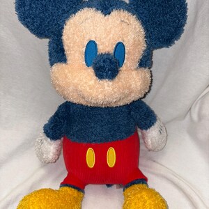 May include: A plush Mickey Mouse toy with a blue head and ears, a tan face, and blue eyes. It wears a blue shirt, red shorts with yellow buttons, and yellow feet. The toy is soft and cuddly.