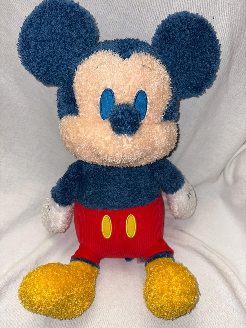 May include: A plush Mickey Mouse toy with a blue head and ears, a tan face, and blue eyes. It wears a blue shirt, red shorts with yellow buttons, and yellow feet. The toy is soft and cuddly.