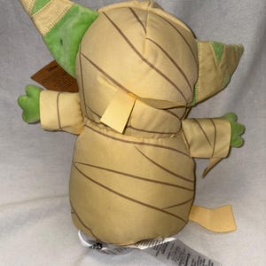 Puede incluir: Peluche de un personaje de la franquicia Star Wars, con un cuerpo beige y rayas marrones, orejas y manos verdes. El juguete tiene una etiqueta adjunta. El dise&ntilde;o recuerda a una momia.