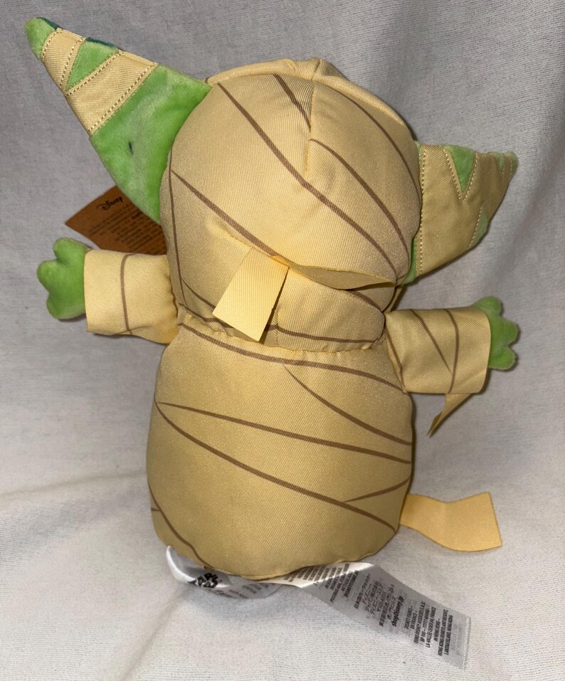 Puede incluir: Peluche de un personaje de la franquicia Star Wars, con un cuerpo beige y rayas marrones, orejas y manos verdes. El juguete tiene una etiqueta adjunta. El dise&ntilde;o recuerda a una momia.