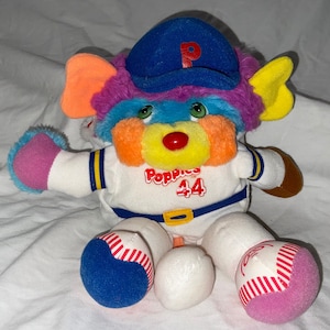 May include: A colorful plush toy wearing a blue baseball cap with a white 'P' on it. The toy is wearing a white baseball uniform with blue and yellow trim. The toy has a red nose and is holding a brown baseball glove. The toy is sitting on a white surface. The toy's uniform has the text 'Popples 44' on the front.