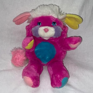 May include: A pink plush toy with white fur on its head, blue eyes, and yellow ears. The toy has a blue circle on its belly and pink and blue accents on its feet.