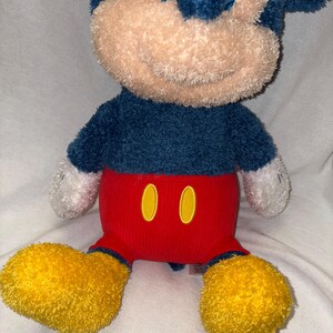 May include: A plush Mickey Mouse toy with a blue body, red pants, and yellow shoes. The toy has a tan face and white hands. The red pants have two yellow oval shapes. The toy is sitting on a white surface.