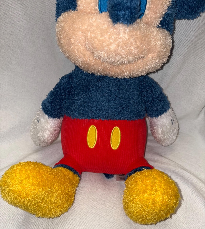 May include: A plush Mickey Mouse toy with a blue body, red pants, and yellow shoes. The toy has a tan face and white hands. The red pants have two yellow oval shapes. The toy is sitting on a white surface.