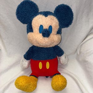 May include: A plush Mickey Mouse toy. The toy features a navy blue head and ears, a tan face, and blue eyes. It wears red shorts with yellow buttons, white gloves, and yellow shoes. The plush toy is soft and cuddly.