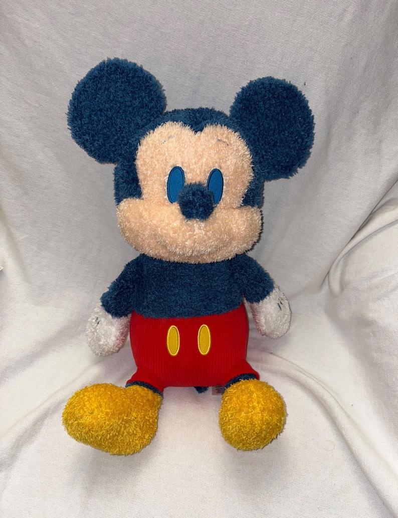 May include: A plush Mickey Mouse toy. The toy features a navy blue head and ears, a tan face, and blue eyes. It wears red shorts with yellow buttons, white gloves, and yellow shoes. The plush toy is soft and cuddly.
