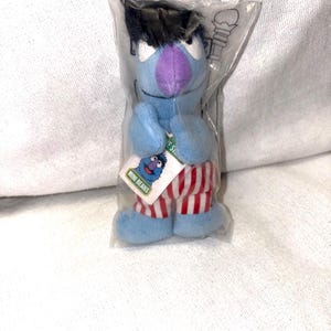 May include: A blue plush toy of a Sesame Street character, packaged in a clear plastic bag. The toy has black hair, a purple nose, and red and white striped shorts. A tag reads "Mini Beans".