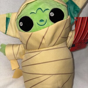 Puede incluir: Un peluche de un personaje de piel verde con grandes ojos negros y una expresi&oacute;n sonriente. El personaje est&aacute; envuelto en tela beige con rayas marrones, que se asemeja a una momia. El juguete tiene una etiqueta de Star Wars.
