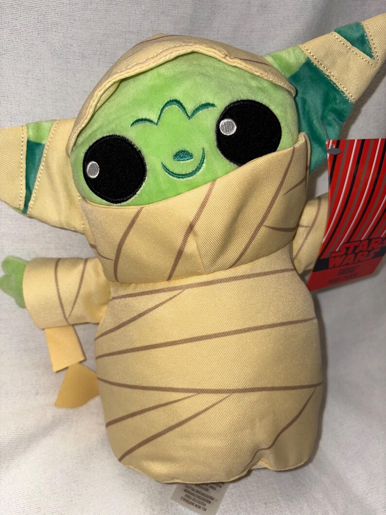 Puede incluir: Un peluche de un personaje de piel verde con grandes ojos negros y una expresi&oacute;n sonriente. El personaje est&aacute; envuelto en tela beige con rayas marrones, que se asemeja a una momia. El juguete tiene una etiqueta de Star Wars.