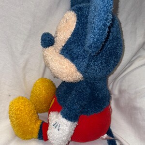 May include: A plush Mickey Mouse toy in a seated position. The toy features a blue head and ears, a tan face, red shorts, yellow shoes, and white gloves. The toy is made of soft, textured fabric.