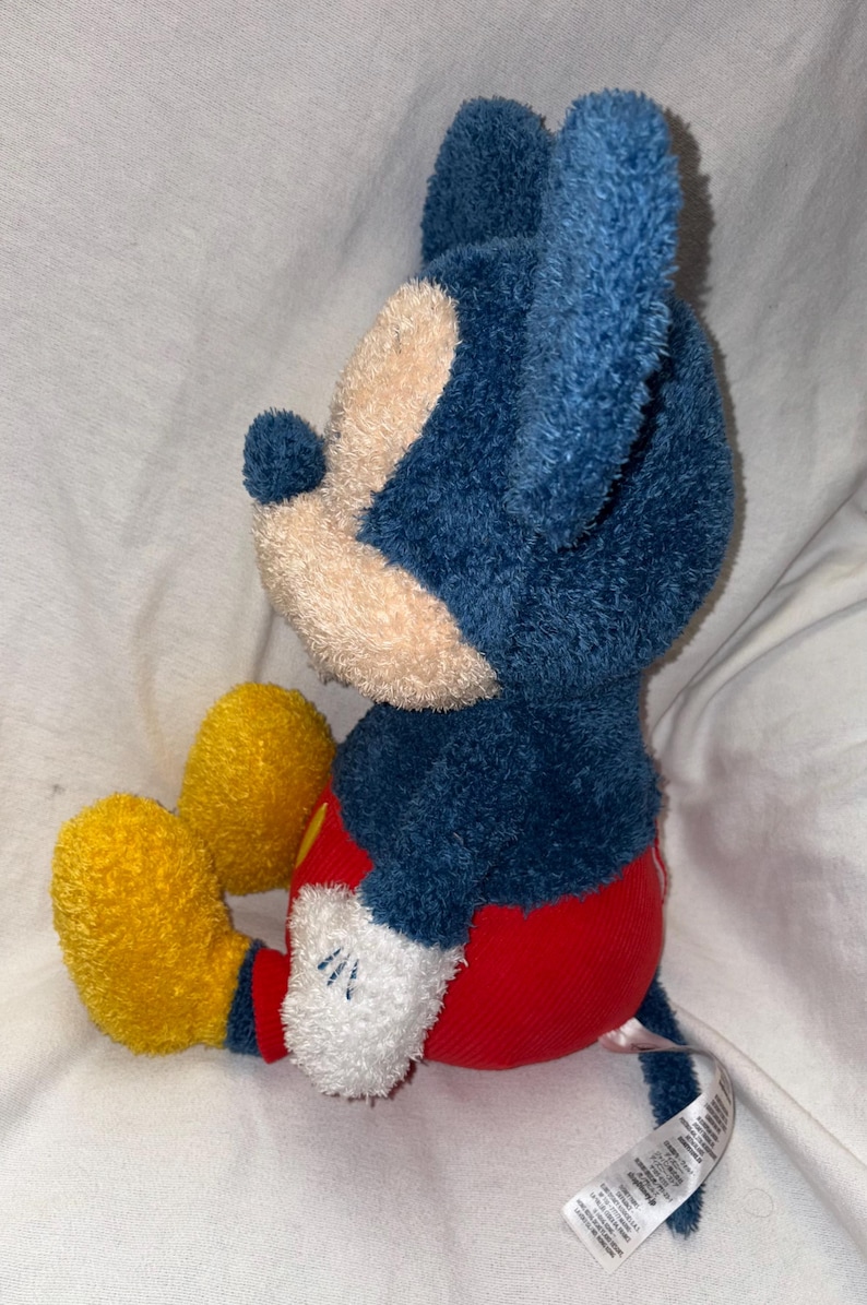 May include: A plush Mickey Mouse toy in a seated position. The toy features a blue head and ears, a tan face, red shorts, yellow shoes, and white gloves. The toy is made of soft, textured fabric.