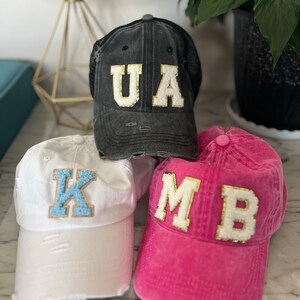 Puede incluir: Tres gorras de béisbol con iniciales. La primera es una gorra negra desgastada con las letras "UA" en dorado y blanco. La segunda es blanca con una "K" azul claro. La tercera es rosa con "MB".