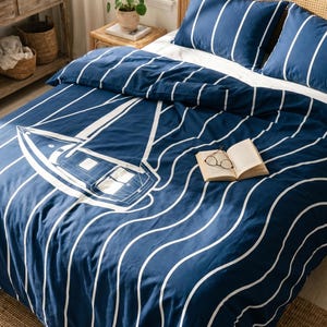 May include: A navy blue duvet cover and two matching pillowcases featuring a white graphic of a sailboat and wavy vertical lines. An open book with a pair of glasses resting on its pages is placed on the centre of the bedspread.