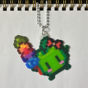May include: A pixelated green creature charm with a rainbow horn and pink cheeks, attached to a white beaded chain. The charm is made of colorful plastic beads, and the background is a white notebook.