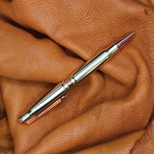 May include: A silver and copper pen made from a repurposed bullet casing. The pen is lying on a brown leather surface.