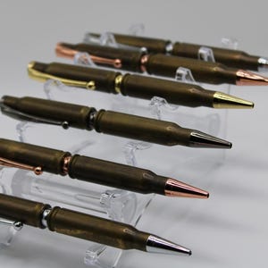May include: A collection of handcrafted pens with a unique design. The pens are made from brass bullet casings and feature various metallic accents, including gold, copper, and silver. The pens are displayed on clear acrylic stands.