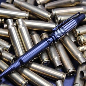 May include: A black tactical pen with a silver American flag design, lying on a bed of spent brass casings.