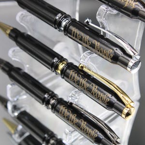 May include: Close-up of several black pens with gold and silver accents. Each pen has the words "We the People" engraved on the barrel. The pens are displayed on a clear, angled stand.