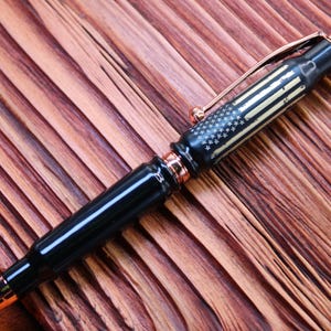 May include: A black and rose gold pen with a bullet casing design. The pen has a white and gold American flag design on the barrel.